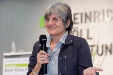 Person mit Mikrofon in der Hand lächelt an der Kameralinse vorbei, im Hintergrund Schriftzug "Heinrich Böll Stiftung" und ein Rednerpult.
