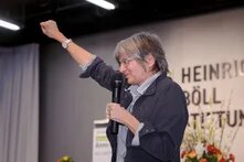 Person mit grauen Haaren und Brille hält ein Mikrofon in der Hand und streckt einen Arm kraftvoll zur Faust geballt in die Höhe. Im Hintegrund Blumen und ein Rednerpult.