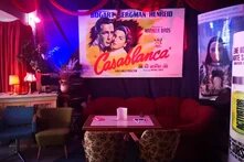 Sitzecke mit Sofa aus rotem Samt sowie Sessel im Foyer eines Kinos, an der Wand geradezu ein Plakat des Films "Casablanca", an der rechten Wand weitere Filmplakate.