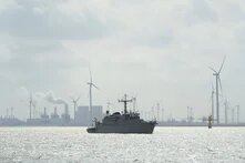 Schiff auf ruhigem Wasser, dahinter Windräder und Industrieanlagen; aus einem Schornstein steigt Rauch auf.