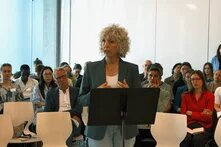 Frau mit Locken spricht an Rednerpult vor sitzendem Publikum in hellem Raum.