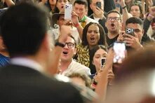 Im (unscharfen) Vordergrund steht eine Person mit dem Rücken zur Kamera, im Hintergrund eine gestikulierende Menschengruppe, die die Person im Vordergrund fotografiert.