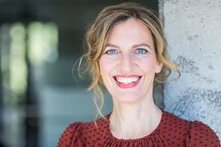 Person mit lockig zurückgesteckten Haaren lehnt an einer Betonwand und lächelt direkt in die Kamera. Nahaufnahme von Gesicht und Oberkörper, helle Augen, verschwommener Innenraum im Hintergrund.