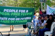Menschen bei Kundgebung im Park. Großes Banner: „Menschen für eine Zukunft ohne fossile Energien“; mehrere Personen sitzen daneben.