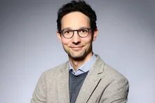 Lächelnde Person mit Brille blickt in die Kamera, Arme verschränkt, vor neutralem Hintergrund.