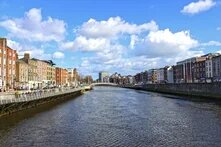 Blick über einen Fluss mit Uferpromenaden, Brücke in der Ferne und mehrstöckigen Häusern auf beiden Seiten unter wolkigem Himmel.