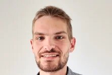 Lächelnde Person mit kurzem Haar und Bart blickt in die Kamera, vor heller Wand.