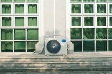 Air conditioning unit in front of a building facade between large windows with curtains; steps run along the front.