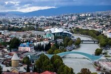 Blick über Tiflis mit Fluss, Fußgängerbrücke, markantem Kuppelbau, dicht bebauter Stadt und Bergen im Hintergrund.