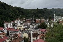A view of a small town with mosques and a church, surrounded by wooded hills under a heavily overcast sky.