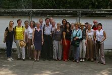 Etwa 20 Personen stehen dicht beisammen vor einer niedrigen Mauer mit Bäumen im Hintergrund und posieren für ein Gruppenfoto.
