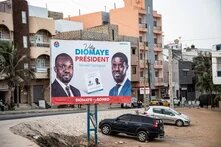Wahlplakat mit Bassirou Diomaye Faye und Ousmane Sonko vor Wohnhäusern; davor Straße, parkende Autos und offener Entwässerungsgraben.