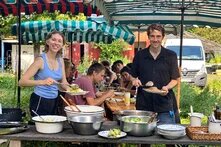 Zwei Personen stehen mit Tellern an einem Buffet im Freien; dahinter sitzen weitere Menschen unter gestreiften Sonnenschirmen.