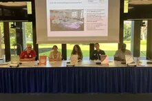 Fünf Personen sitzen an einem Konferenztisch. Dahinter zeigt eine Leinwand eine Präsentation zu Zielen der Entwicklungszusammenarbeit.