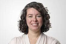 Eine lächelnde Frau mit schulterlangen, dunklen Locken steht mit verschränkten Armen vor einem hellen Hintergrund.