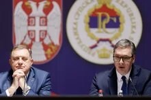 Two politicians sit at a press conference in front of Serbian state symbols. The man on the right speaks while the man on the left listens with his eyes closed.