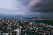 Blick über dicht bebaute Stadtviertel Istanbuls auf den Bosporus und eine beleuchtete Brücke. Darüber türmen sich dunkle Gewitterwolken.