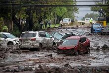Mehrere beschädigte Autos stehen auf einer von Schlamm, Steinen und Ästen bedeckten Straße. Im Hintergrund stehen Menschen und Einsatzkräfte.