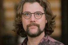 Ein Mann mit schulterlangen, welligen Haaren, Bart und runder Brille blickt direkt in die Kamera. Der Hintergrund ist unscharf.