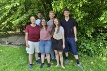 Sechs Menschen stehen für ein Gruppenfoto auf einer Wiese vor Bäumen.