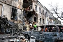 Ein schwer beschädigtes Gebäude mit eingestürzten Wänden. Davor stehen ein ausgebranntes Auto und Einsatzkräfte zwischen Trümmern.