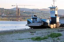 A ferry on a sandy riverbank; in the background, a bridge with a crane and stay cables is under construction.