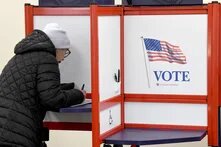 Eine Frau füllt in einer Wahlkabine einen Stimmzettel aus. An der Trennwand sind die US-Flagge und das Wort „VOTE“ (Wählen) zu sehen.
