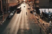 Foto: Breite Stadtstraße in einem Geschäftsviertel bei Sonnenuntergang. Ein Bus und Autos fahren, zwei Fußgänger*innen überqueren die Straße. Hochhäuser im Hintergrund.