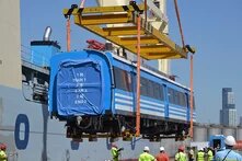 Foto: Ein blau-weißer Eisenbahnwagen mit chinesischen Schriftzeichen wird per Kran von einem Schiff verladen. Arbeiter*innen in Warnwesten und Helmen stehen unten. Hochhäuser im Hintergrund.