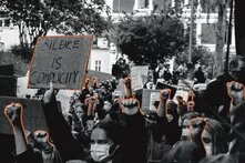 Photo: A large protest with people raising their fists and holding signs. Some signs read "SILENCE IS VIOELENCE" and "SILENCE IS COMPLICITY." The crowd is dense.