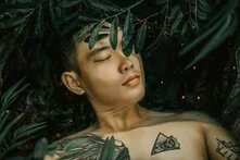 A shirtless person with short dark hair and closed eyes lies among dark green leaves. Visible tattoos include a stylized wing, a triangle, and text.