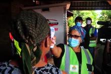 Eine Person in Sicherheitsweste misst kontaktlos die Temperatur einer anderen Person. Weitere Personen mit Masken im Hintergrund.