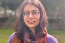  Eine Frau mit Brille und langen, braunen Haaren steht draußen im Sonnenschein. Sie trägt einen lilafarbenen Pullover und einen braunen Schal. Der Hintergrund ist unscharf mit Bäumen und Rasen.