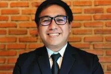 Photo: A person with glasses and a tie in a dark suit, smiling in front of a brick wall.
