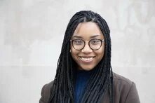 Person mit langen Zöpfen und Brille lächelt, Blick zur Kamera, neutraler Hintergrund.