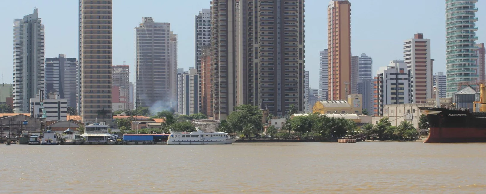 Mehrere Hochhäuser hinter Flussufer mit Boot und Häusern im Vordergrund, Rauch steigt zwischen Bäumen auf.