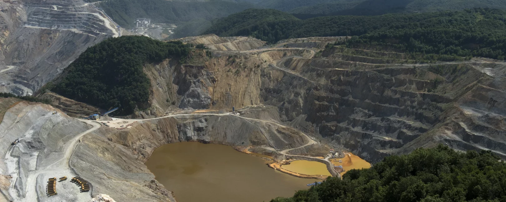 Foto: Weitläufiger Tagebau in hügeliger, bewaldeter Landschaft. Ein großer brauner See ist in der Mitte der Grube sichtbar.