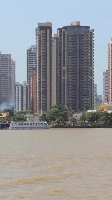 Several high-rise buildings behind the riverbank with a boat and houses in the foreground, smoke rising between the trees.