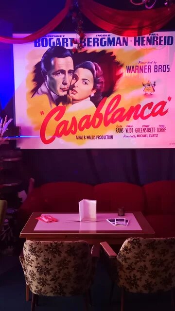 Sitzecke mit Sofa aus rotem Samt sowie Sessel im Foyer eines Kinos, an der Wand geradezu ein Plakat des Films "Casablanca", an der rechten Wand weitere Filmplakate.