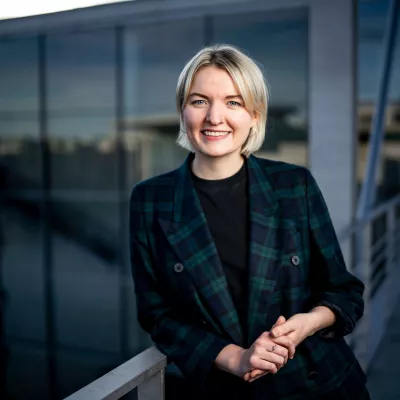 Porträt Leonie Hopgood (blonde Haare, grün-blau-kariertes Sakko, lehnt an einem Geländer, lächelnd)