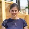 Antje Gust steht vor einer Wand aus Holz. Sie trägt ein blaues T-Shirt, eine lange Kette und hat ihre braunen Haare in einem Pferdeschwanz.