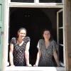 Zwei Frauen stehen in einem Haus und schauen durch ein offenes Fenster nach draußen. Beiden tragen ein gemustertes Kleid und ihre Haare in einem Zopf.