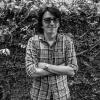 Portrait of Bill Barreto. Black and white photo. He is wearing sunglasses, has his arms crossed, and is standing in front of a hedge.