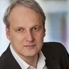 Portrait Marc Buggeln, Person mit graumeliertem Haar, hoher Stirn, im Anzug