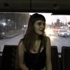 A young person with long, wavy brown hair and straight bangs sits on a bus. Through the window, a busy street, car headlights, and a sunset are visible.