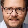Foto: Porträt einer Person mit Brille und Bart vor neutralem Hintergrund, Blick direkt in die Kamera.