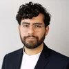 Cristian González Cabrera Person mit Brille, Bart und lockigem Haar blickt ernst in die Kamera, vor hellem Hintergrund.