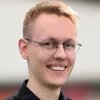 Eine lächelnde Person mit Brille blickt in die Kamera, kurzer Haarschnitt, unscharfer heller Hintergrund.