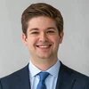 Ein junger Mann mit kurzen, gestylten Haaren lächelt und blickt frontal in die Kamera. Er trägt Anzug, Hemd und Krawatte.