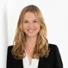 Eine Person mit langen, welligen Haaren steht vor hellem Hintergrund und lächelt in die Kamera.